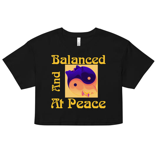 AS Colour brand, product 4062, women's crop tee. Black top with yellow quote "Balanced And At Peace" with bluish/purple and yellowish/pinkish/peachish yin-yang heads image. Front of the shirt shown on white background. JAMILLIAH'S WISDOM IS TIMELESS SHOP - wisdomistimeless.com.