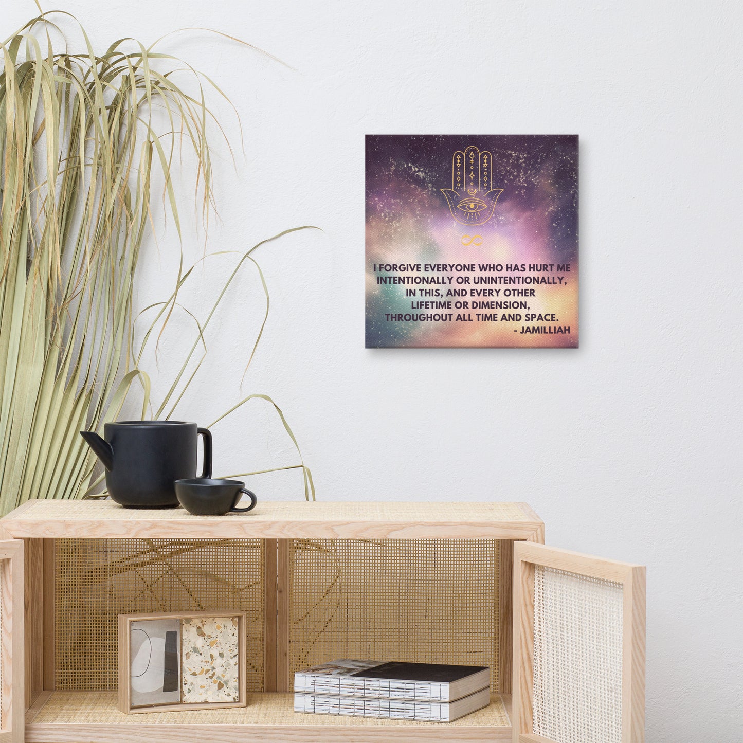 16x16 inch, semi-gloss, textured canvas print wall art. Purple/green/yellow space/universe/cosmos design with spiritual hamsa hand/infinity symbol/sign brand logo image, and original wise quote mantra/saying/tagline/motto/slogan. "I Forgive Everyone Who Has Hurt Me Intentionally Or Unintentionally, In This, And Every Other Lifetime Or Dimension, Throughout All Time And Space. - Jamilliah." Shown on wall next to plant, cabinet, mug, and teapot. JAMILLIAH'S WISDOM IS TIMELESS SHOP - wisdomistimeless.com.