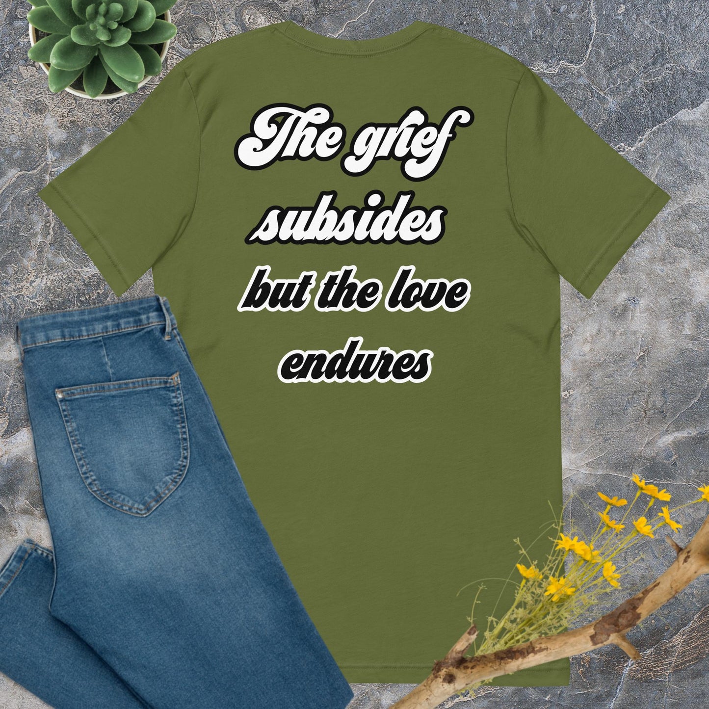 Bella + Canvas 3001 t-shirt. Olive green colored, back view. White and black scripted written quote "The grief subsides but the love endures." Shown laying flat on grey marble with blue jeans, a green succulent plant, and a brown twig with yellow flowers. Jamilliah's Wisdom Is Timeless Shop - wisdomistimeless.com.