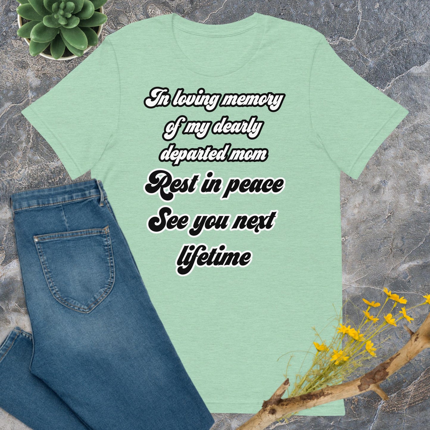 Bella + Canvas 3001 t-shirt. Heather prism mint colored (light green), front view. White and black scripted written quote "In loving memory of my dearly departed mom. Rest in peace. See you next lifetime." Shown laying flat on grey marble with blue jeans, a green succulent plant, and a brown twig with yellow flowers. Jamilliah's Wisdom Is Timeless Shop - wisdomistimeless.com.