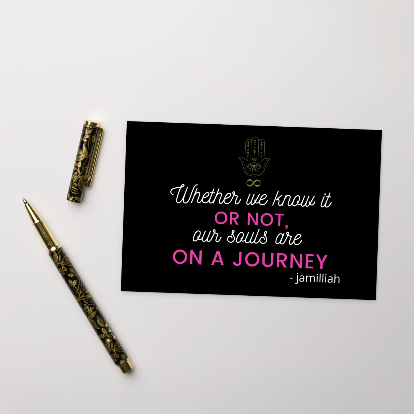 4" x 6", black, glossy, cardboard postcard with pink and white writing, original hamsa hand/infinity symbol brand logo, and wise quote mantra/saying/motto/tagline. "Whether We Know It Or Not, Our Souls Are On A Journey" - Jamilliah. Slogan is "JAMILLIAH'S THE SOUL-PATH JOURNEY PODCAST" tagline. Shown with fancy pen and cap. JAMILLIAH'S WISDOM IS TIMELESS SHOP - wisdomistimeless.com.