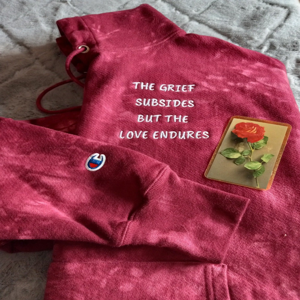 Unisex mulled berry Champion tie-dye hoodie. Gray embroidered quote, "The Grief Subsides But The Love Endures." Shown with rose funeral/memorial card. JAMILLIAH'S WISDOM IS TIMELESS SHOP - wisdomistimeless.com.
