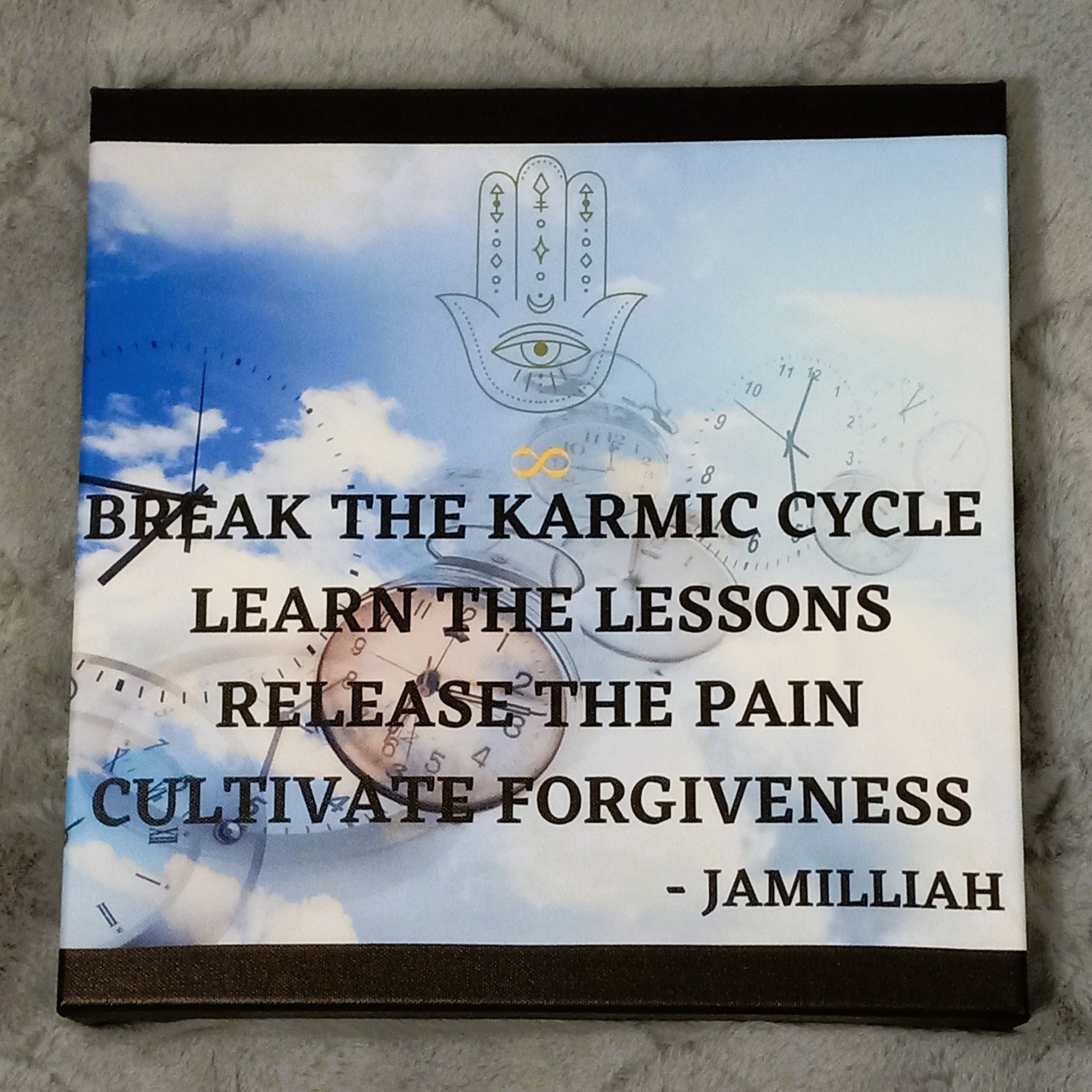 12x12 inch canvas print with original logo and authentic saying. Wall art with clocks in the blue, clouded sky with hamsa hand/infinity sign logo, and black printed, unique, wise quote mantra. "Break The Karmic Cycle, Learn The Lessons, Release The Pain, Cultivate Forgiveness." - Jamilliah - Images surrounding majority of textured canvas, with the remainder of the canvas colored black. Shown on wall above bed. JAMILLIAH'S WISDOM IS TIMELESS SHOP - wisdomistimeless.com.