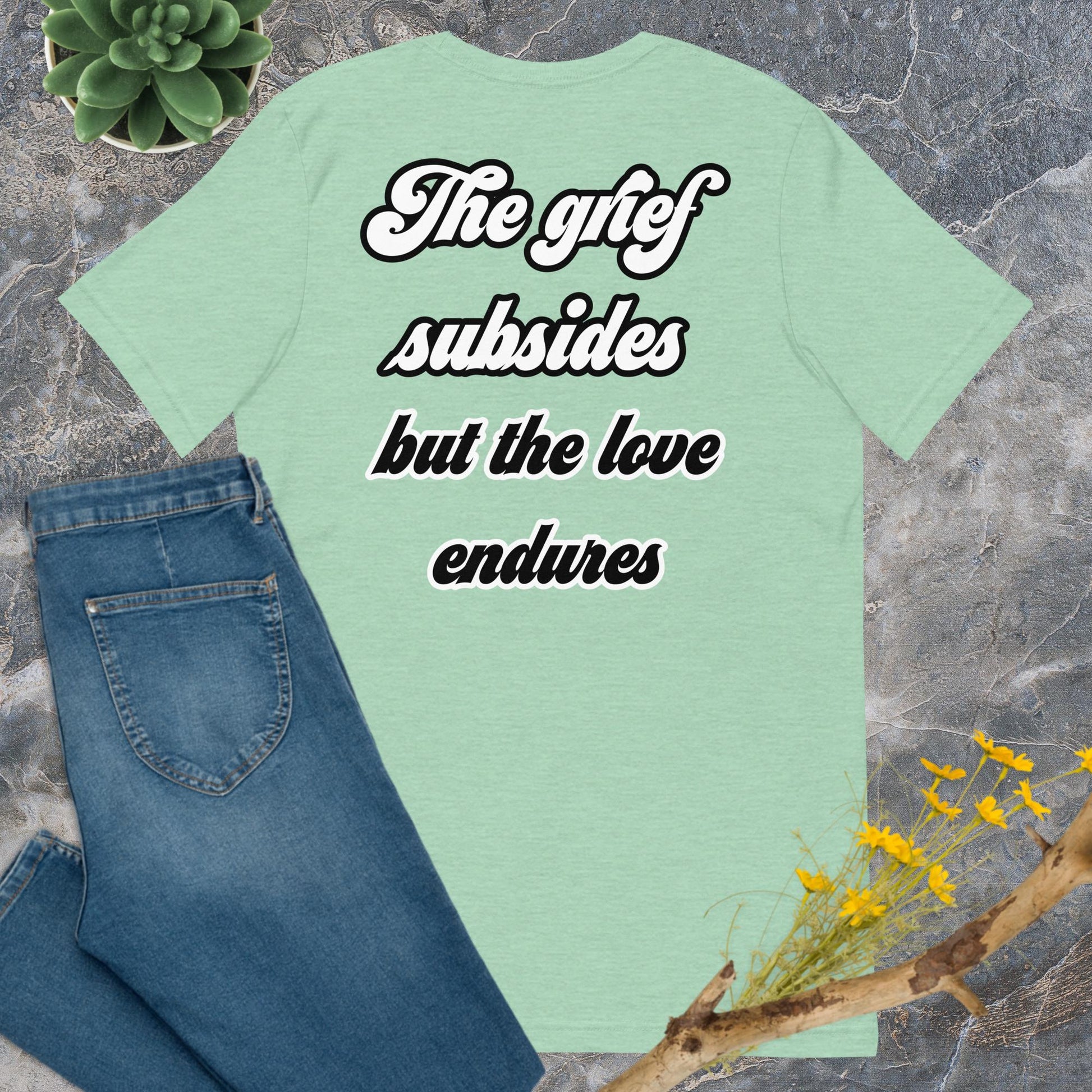 Bella + Canvas 3001 t-shirt. Heather prism mint colored (light green), back view. White and black scripted written quote "The grief subsides but the love endures." Shown laying flat on grey marble with blue jeans, a green succulent plant, and a brown twig with yellow flowers. Jamilliah's Wisdom Is Timeless Shop - wisdomistimeless.com.
