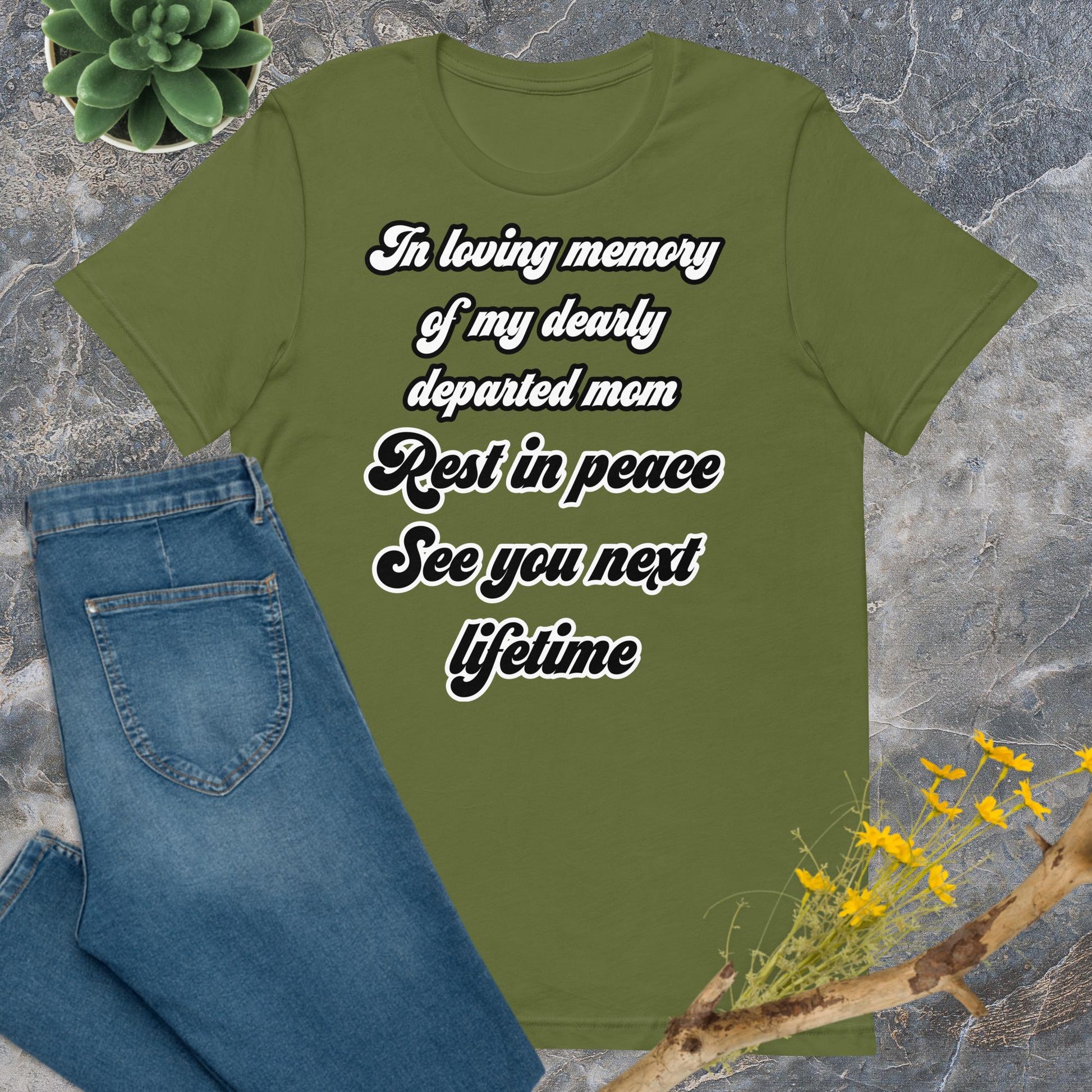 Bella + Canvas 3001 t-shirt. Olive green colored, front view. White and black scripted written quote "In loving memory of my dearly departed mom. Rest in peace. See you next lifetime." Shown laying flat on grey marble with blue jeans, a green succulent plant, and a brown twig with yellow flowers. Jamilliah's Wisdom Is Timeless Shop - wisdomistimeless.com.
