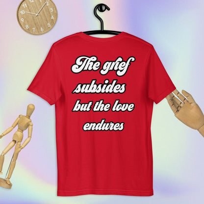 Unisex Bella + Canvas 3001 T-Shirt. Red colored, back view. White and black scripted written quote "The grief subsides but the love endures." Shown on a hanger with lightly colored pink, purple, and yellow background with a wooden clock, wooden figurine, and wooden hand. Sizes SM, M, L, XL, 2X.