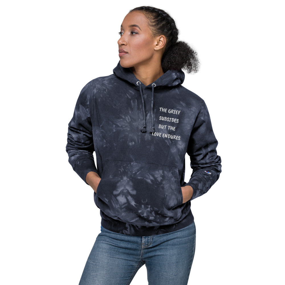 Unisex, navy colored Champion tie-dye hoodie. Embroidered quote, "The Grief Subsides But The Love Endures." Two-ply hood with matching drawcords, front pouch pocket, and embroidered "C" Champion logo on left sleeve. 1×1 rib knit side panels, sleeve cuffs, and bottom hem. Sizes small, medium, large and extra large. Show on female model. JAMILLIAH'S WISDOM IS TIMELESS SHOP - wisdomistimeless.com.