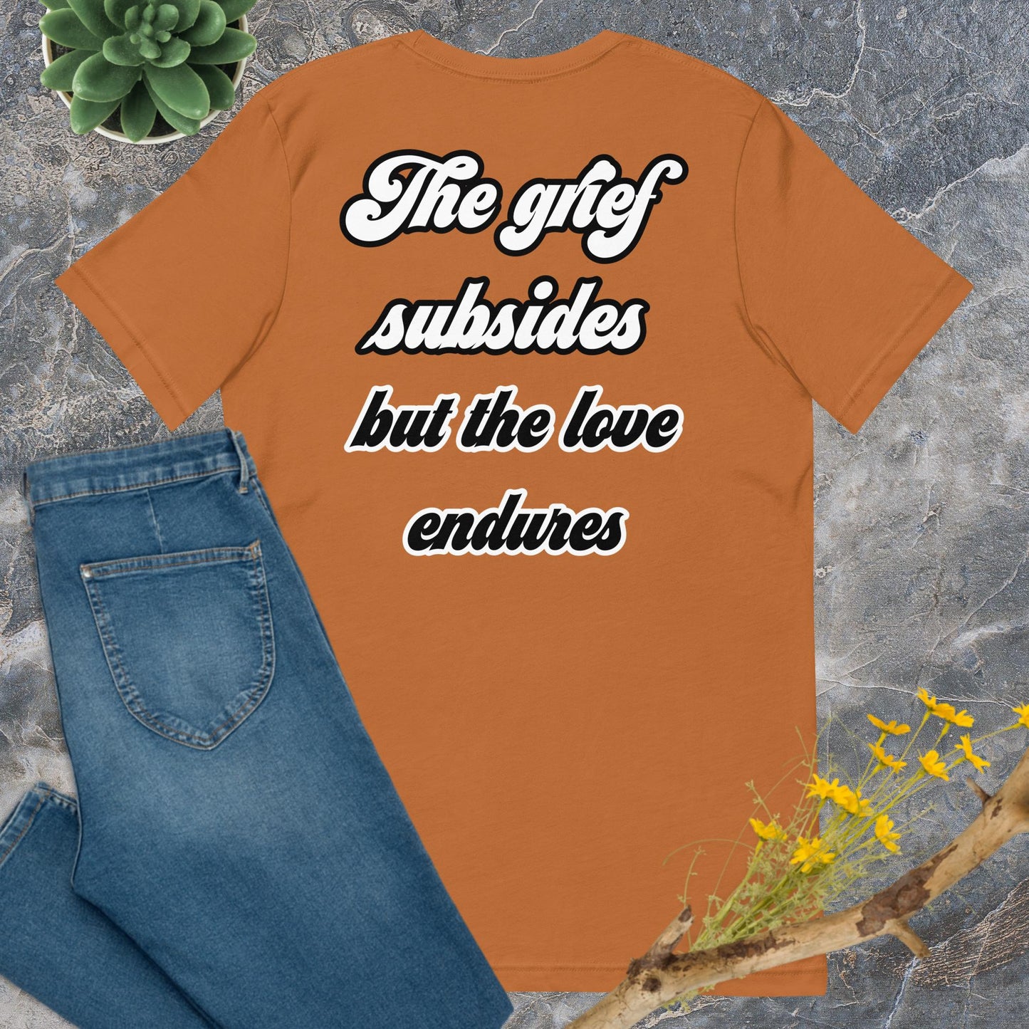 Bella + Canvas 3001 t-shirt. Toast colored (orange/brown), back view. White and black scripted written quote "The grief subsides but the love endures." Shown laying flat on grey marble with blue jeans, a green succulent plant, and a brown twig with yellow flowers. Jamilliah's Wisdom Is Timeless Shop - wisdomistimeless.com.