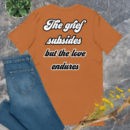 Bella + Canvas 3001 t-shirt. Toast colored (orange/brown), back view. White and black scripted written quote "The grief subsides but the love endures." Shown laying flat on grey marble with blue jeans, a green succulent plant, and a brown twig with yellow flowers. Jamilliah's Wisdom Is Timeless Shop - wisdomistimeless.com.