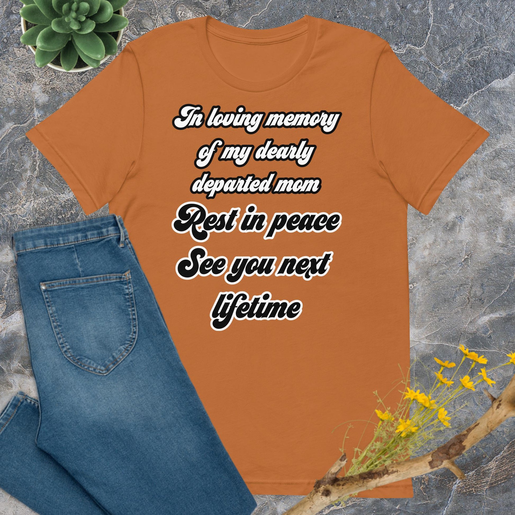 Deceased mom gifts for mother's day. Unisex Bella + Canvas 3001 T-Shirts. Toast colored (orange/brown), front view. White and black scripted written quote "In loving memory of my dearly departed mom. Rest in peace. See you next lifetime."  Shown laying flat on grey marble with blue jeans, a green succulent plant, and a brown twig with yellow flowers. Jamilliah's Wisdom Is Timeless Shop - wisdomistimeless.com.
