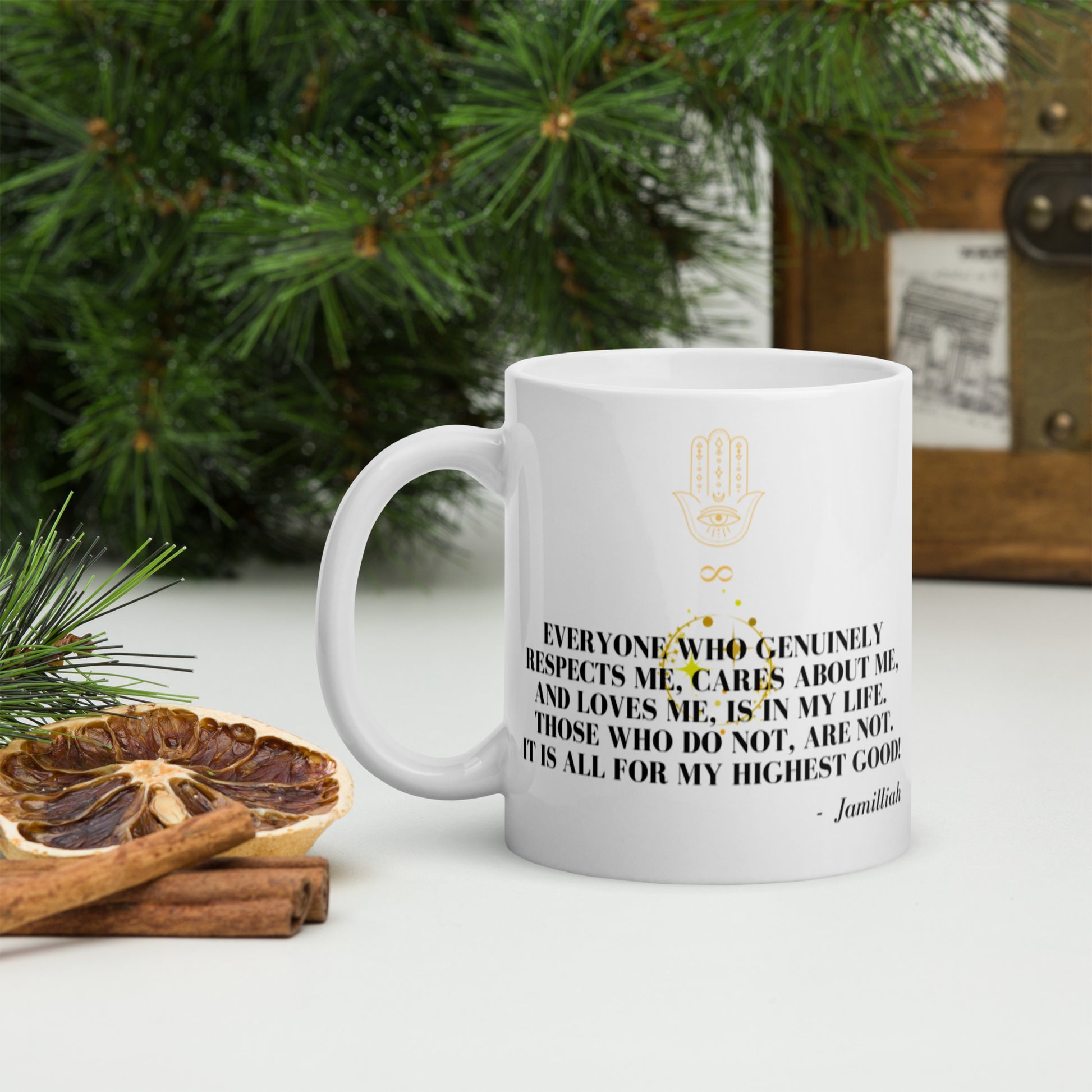 White glossy 11oz mug with original black and yellow design, logo, and inspirational quote, left handle. Spiritual, supernatural hamsa hand/infinity symbol logo. Wise quote mantra. "Everyone Who Genuinely Respects Me, Cares About Me, And Loves Me, Is In My Life. Those Who Do Not, Are Not. It Is All For My Highest Good!" - Jamilliah - Shown on counter with green tree and cinnamon. JAMILLIAH'S WISDOM IS TIMELESS SHOP - wisdomistimeless.com.